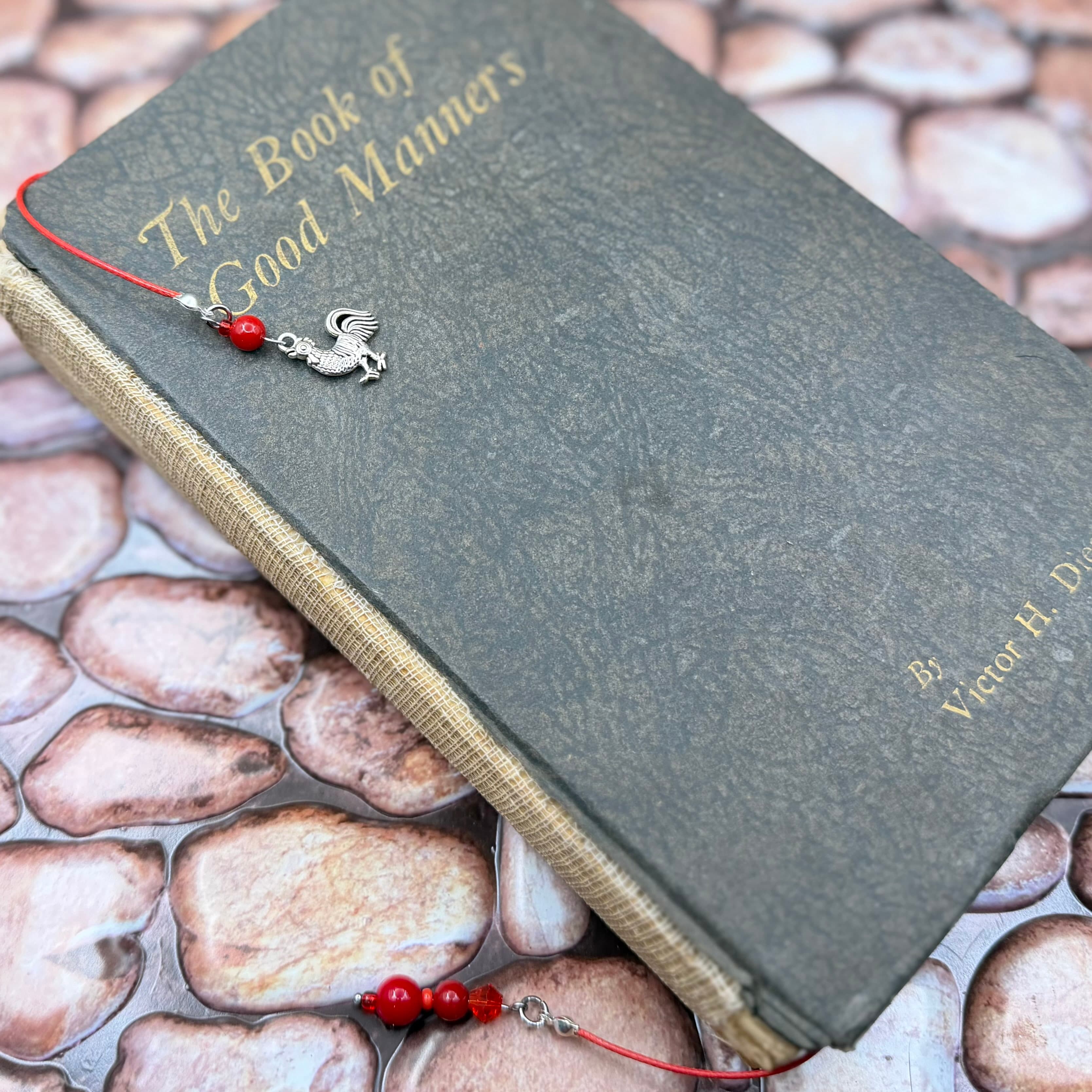 Vintage gray hardcover book titled 'The Book of Good Manners' with gold lettering, featuring a red cord bookmark adorned with a silver rooster charm and red beads.