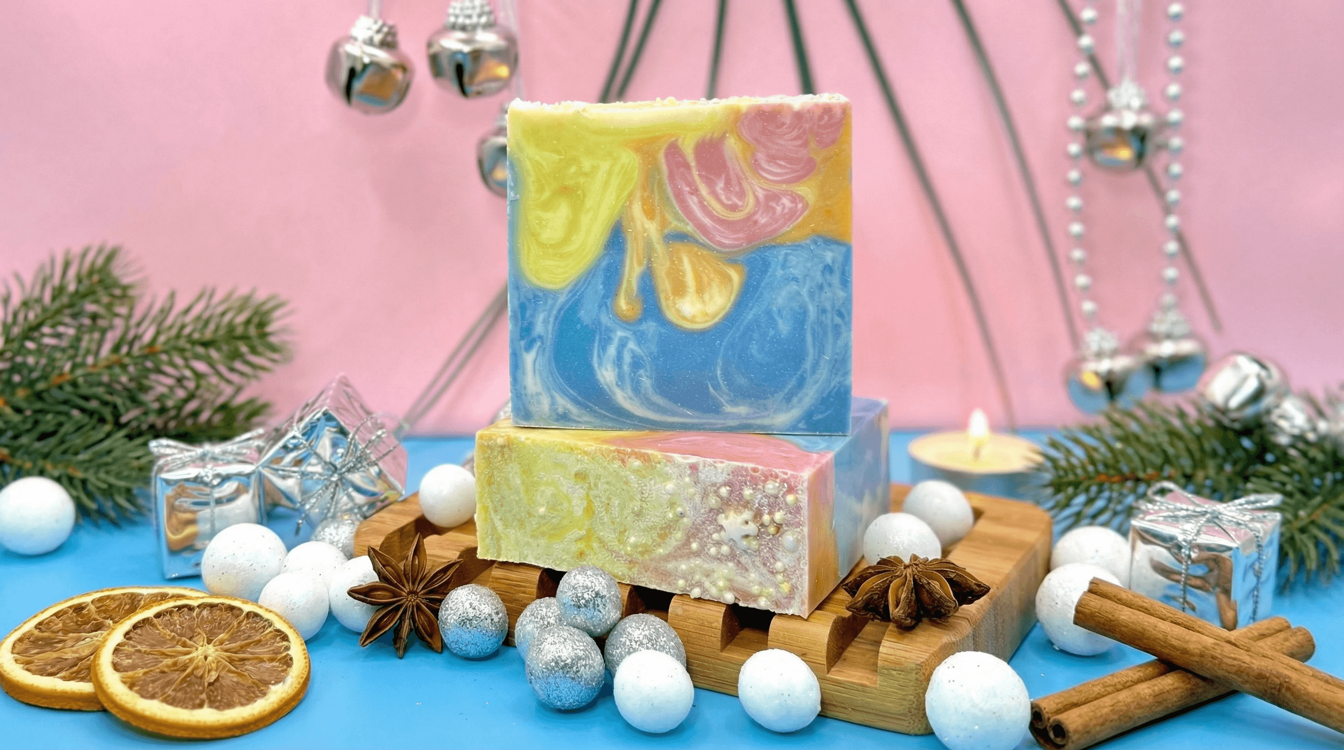 Two marbled soap bars with swirled yellow, pink, and blue layers displayed on a wooden soap dish. Styled with dried citrus slices, cinnamon sticks, star anise, and silver holiday decorations.