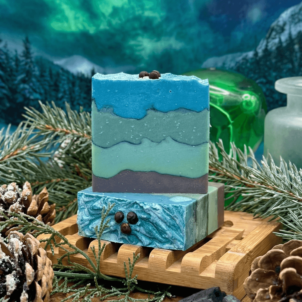 Layered soap bars in ocean-inspired blue, teal, and charcoal stripes with wavy edges, topped with juniper berries. Displayed on bamboo with pine branches, pinecones, and northern lights backdrop.