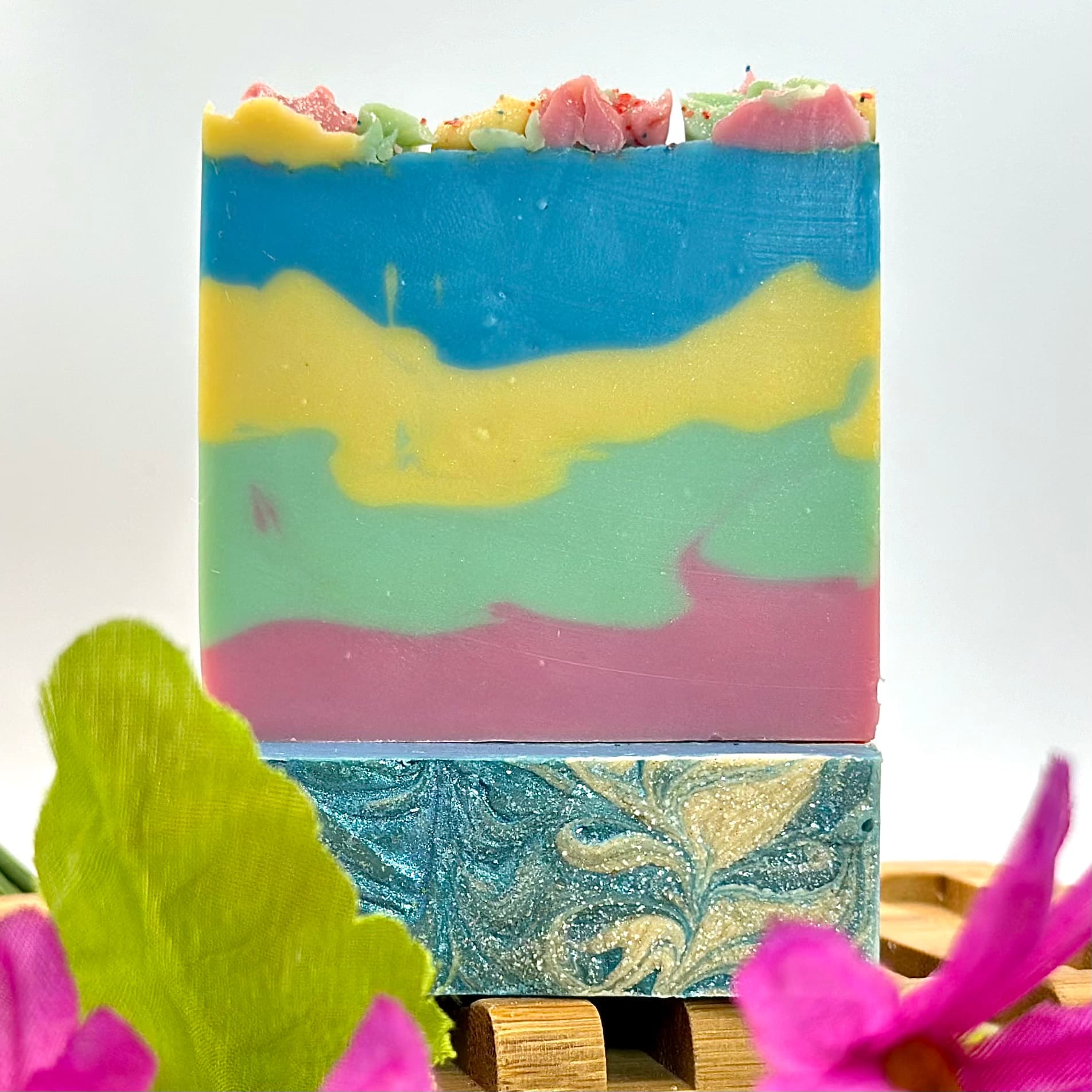 Two stacked handmade soap bars with colorful wavy layers in blue, yellow, pink, and green. Top bar has textured candy-like topping, bottom bar features swirled marbled design.