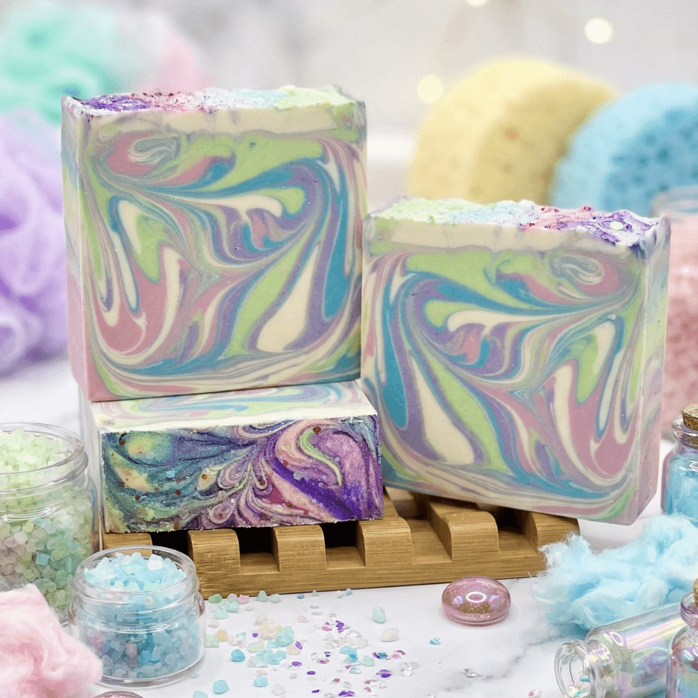 Three rectangular bars of handmade soap with pastel rainbow swirl patterns in purple, pink, green, blue, and cream, topped with purple glitter, displayed on a wooden soap rack.
