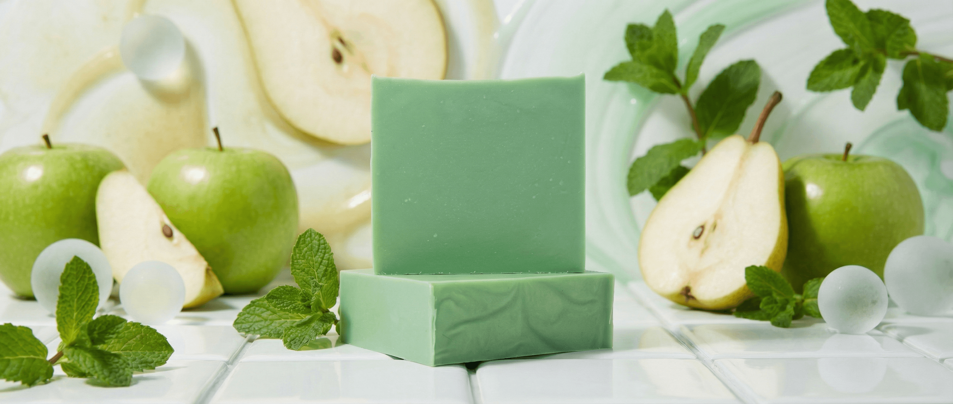 Sage green handcrafted soap bar with smooth matte finish, displayed on matching geometric platform surrounded by fresh green apples, pear slices, and mint leaves on white wood surface.