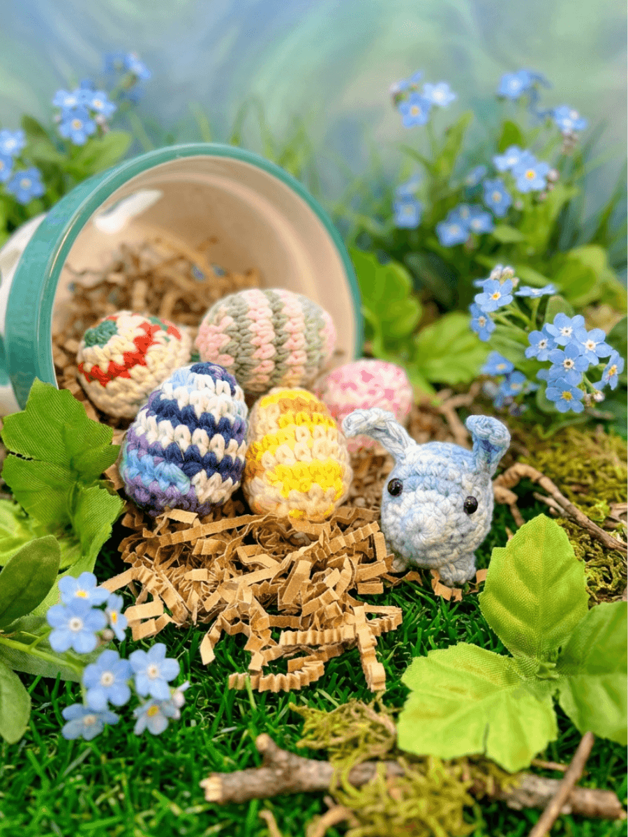 Six colorful crocheted eggs in striped and solid patterns spill from a teal basket onto brown paper shreds, beside a small blue crocheted bunny, surrounded by blue forget-me-not flowers and green moss.