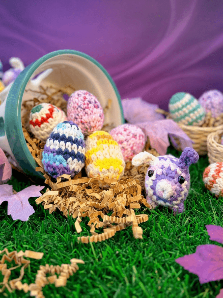Crocheted purple and white bunny with button eyes sits beside colorful striped crocheted eggs spilling from a teal-rimmed basket onto artificial grass with tan paper shred.